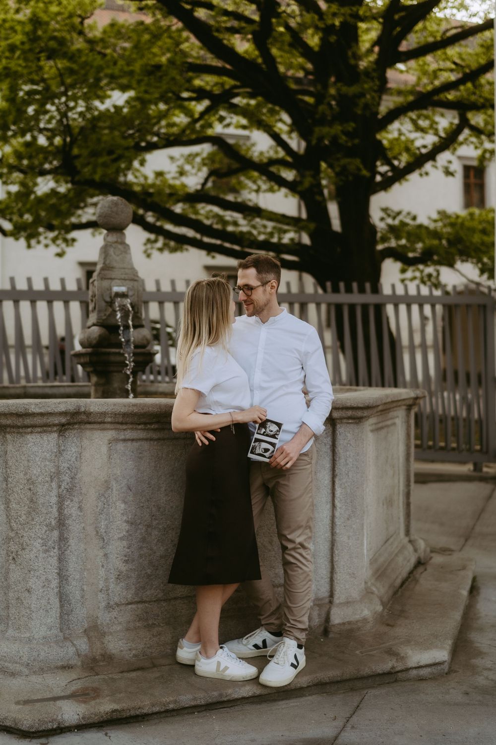 Happy couple holding ultrasound photos by a stone fountain outdoors, surrounded by lush green trees.