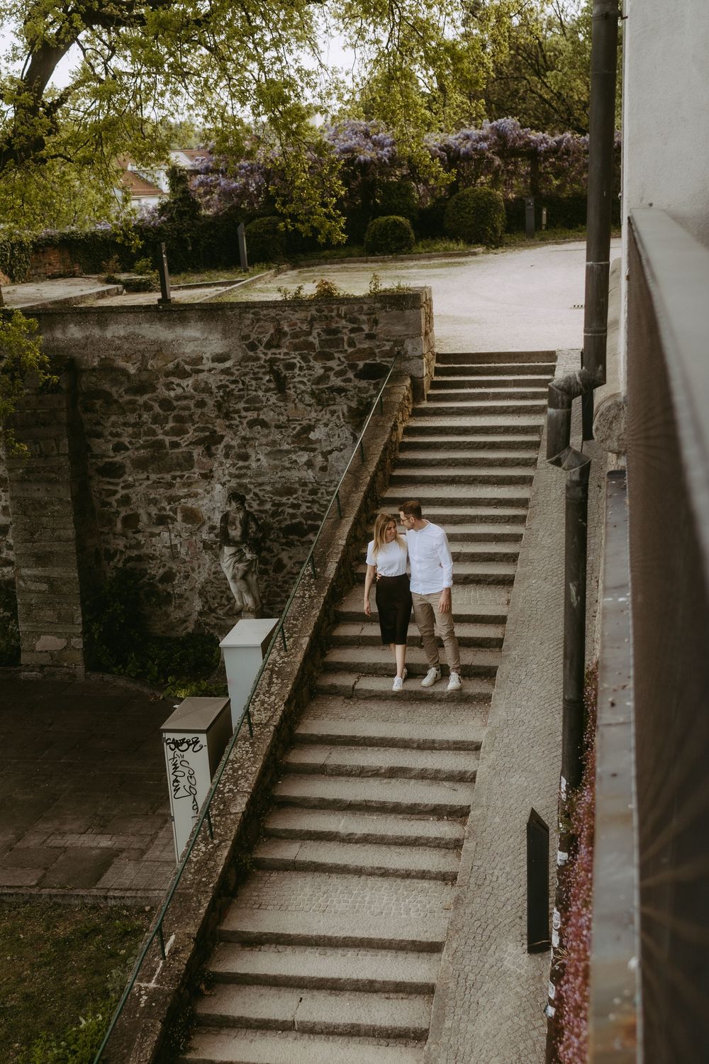 Couple embracing on historic stone staircase with wisteria blooms and cobblestone street in background.