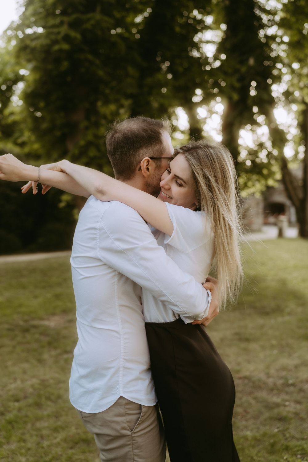 A couple embracing and kissing in a lush green park, woman in black skirt and man in white shirt.