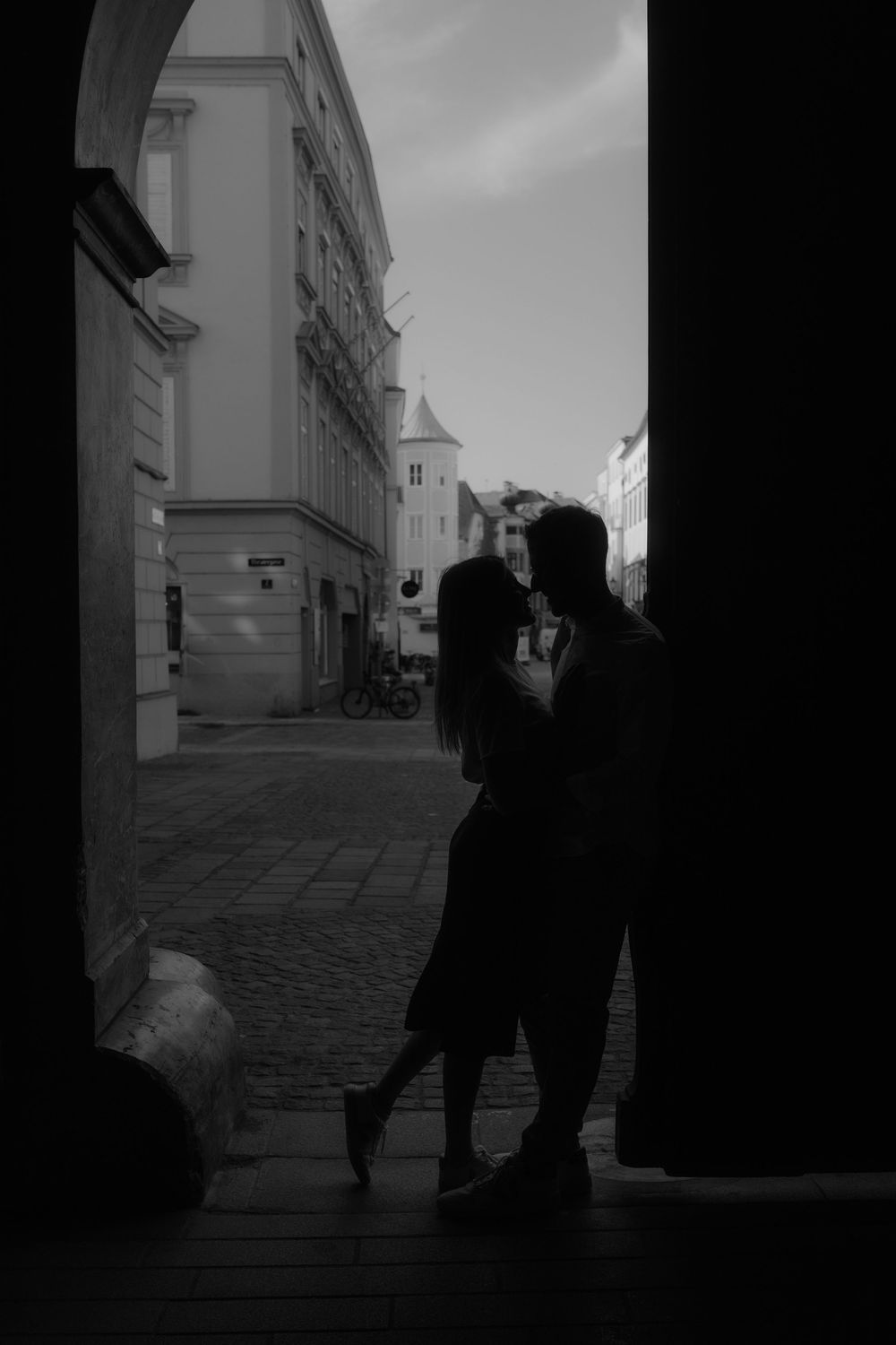 Romantic couple silhouette kissing in a European cobblestone alley framed by dark archway in black and white.
