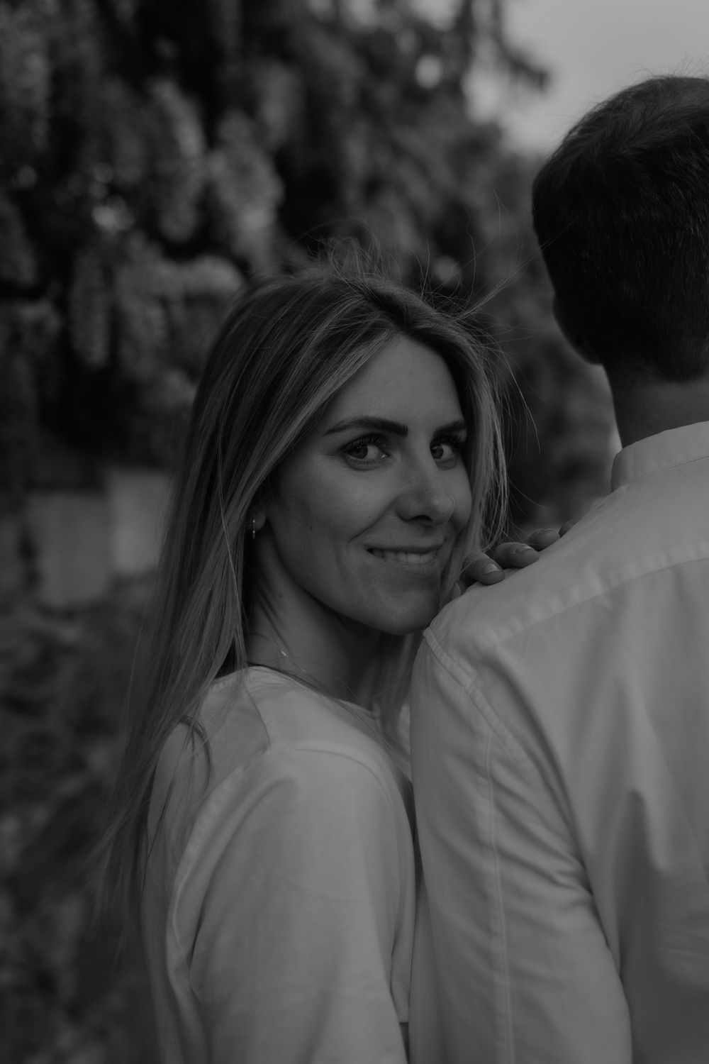 Black and white photo of a smiling blonde woman in white top glancing back, with a man beside her outdoors.