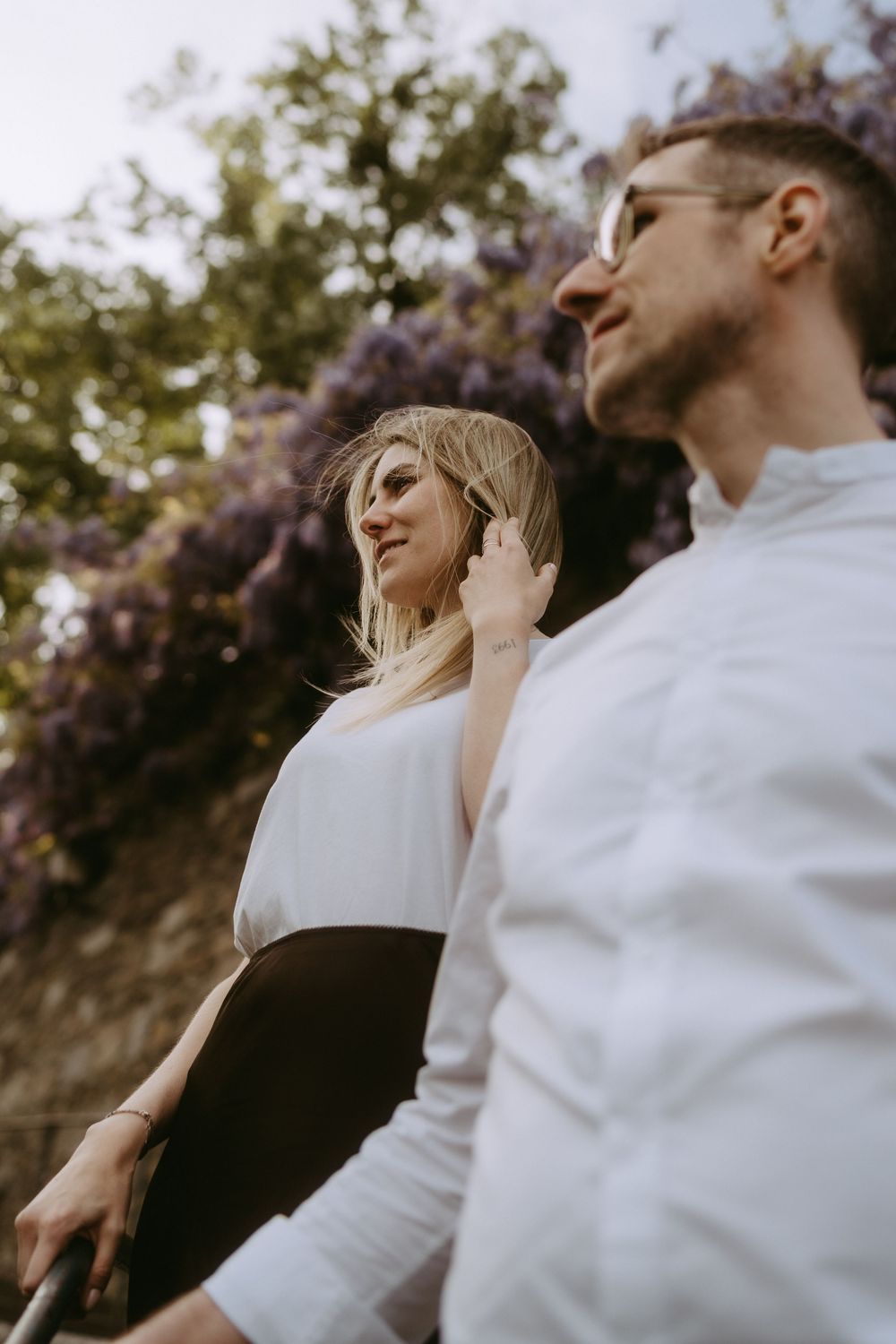 Stylish couple in white shirts posing outdoors near blooming purple wisteria in soft natural light.