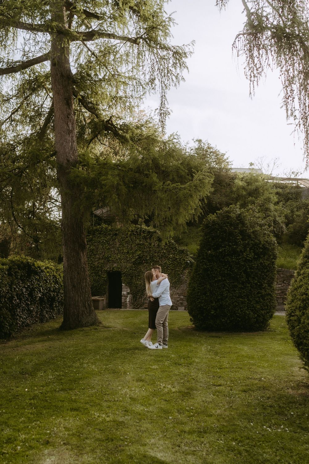 Couple embracing in a lush green garden with tall trees and ivy-covered stone building backdrop.