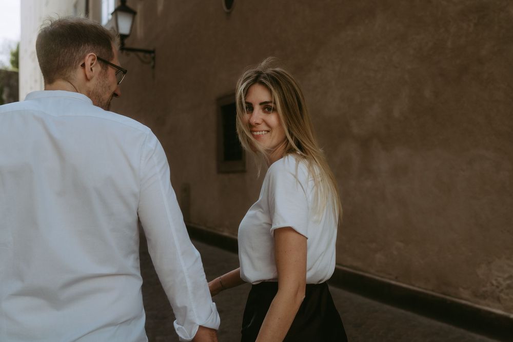 Couple holding hands walking together, woman in white t-shirt smiling over shoulder near rustic wall.
