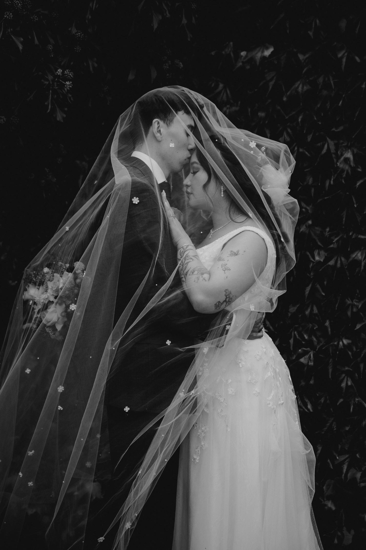 Black and white photo of bride and groom sharing romantic moment under flowing veil outdoors.