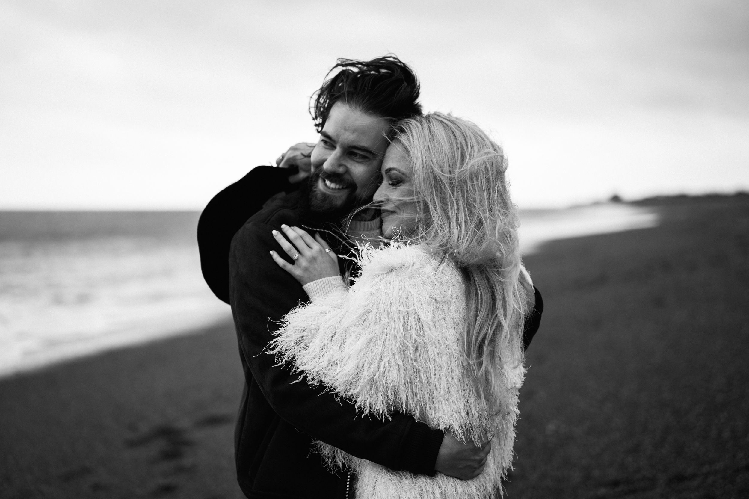 A romantic black and white beach engagement photo session with a couple embracing in flowing white dress.
