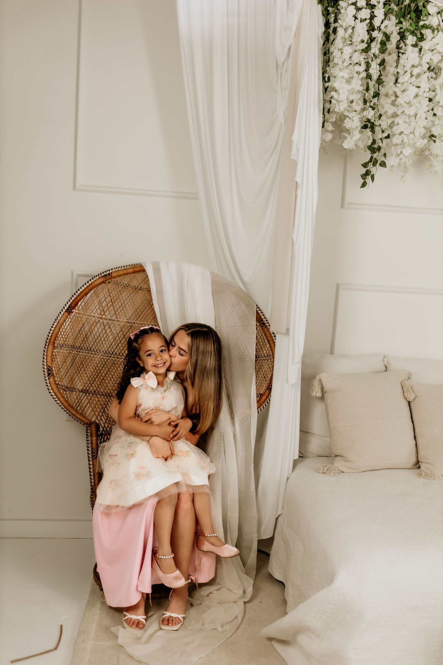 A mother kisses her young daughter while sitting in a wicker chair in a bright white room with floral decor.