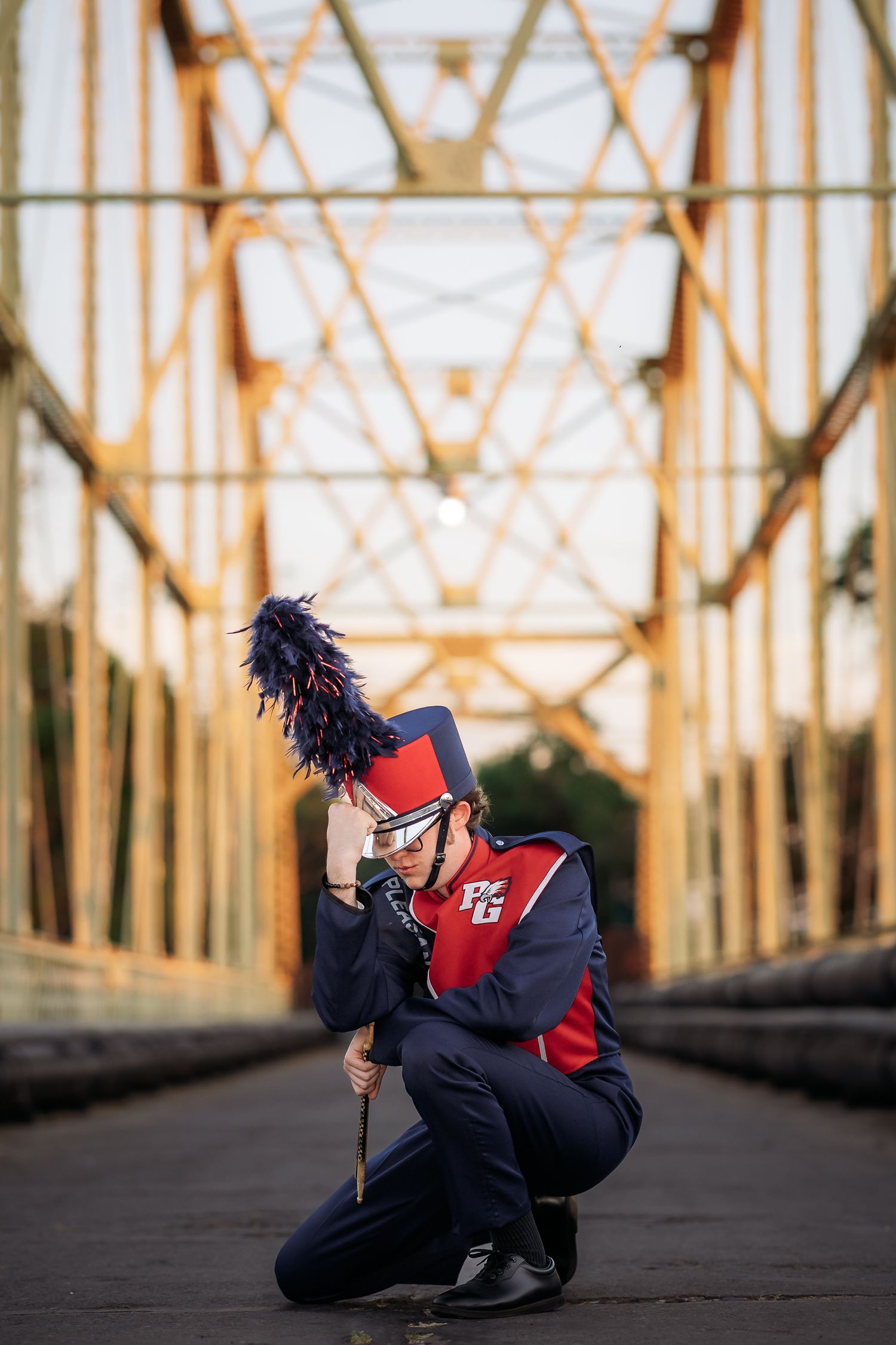 Marching band member in red and blue uniform kneeling on a bridge, holding a baton with a feathered hat.