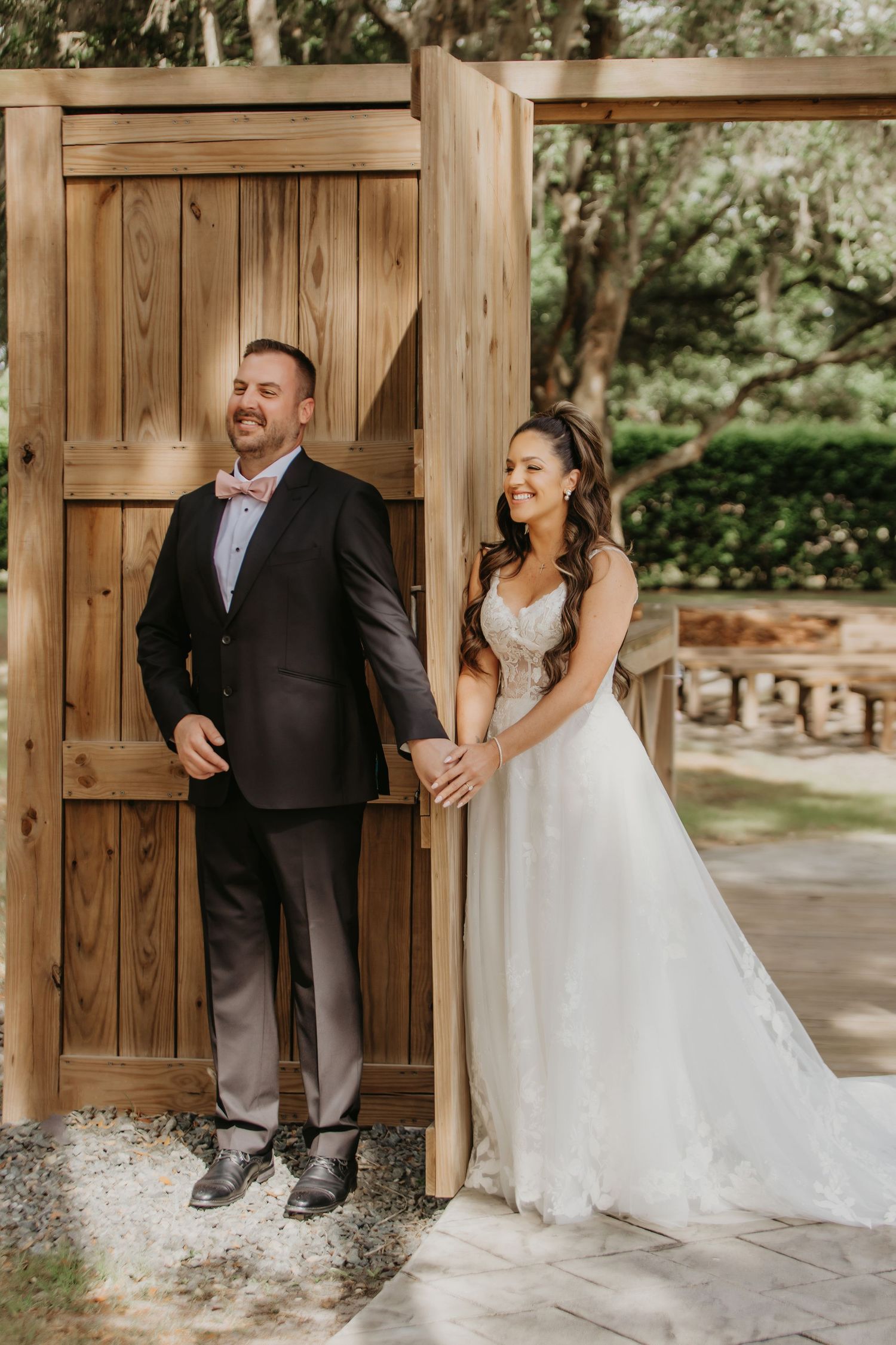 Bride in white gown and groom in dark suit hold hands at wooden door during outdoor wedding first look.