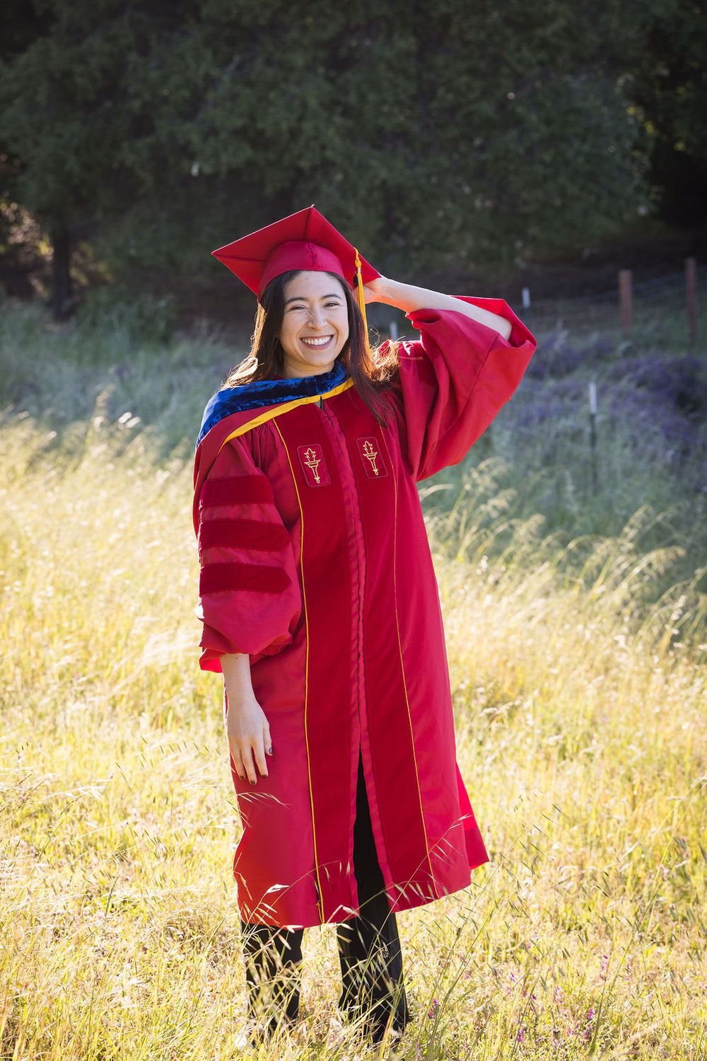 Smiling woman in red graduation cap and gown standing in sunlit field with trees in background.