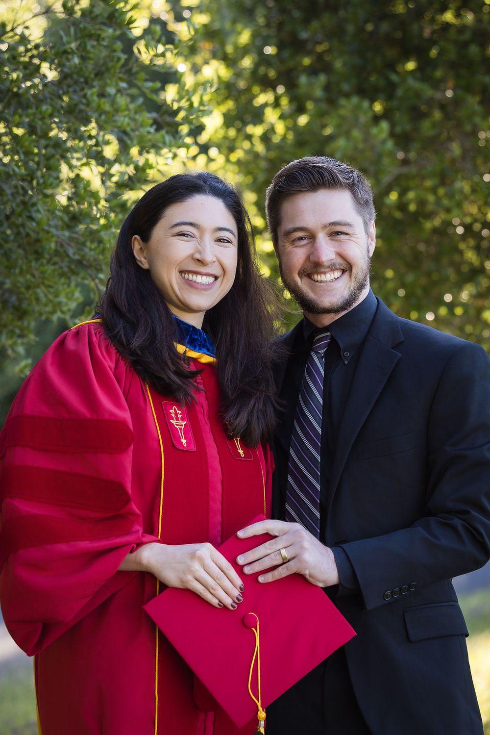 A smiling woman in red graduation robes poses with a man in a black suit outdoors near green trees.