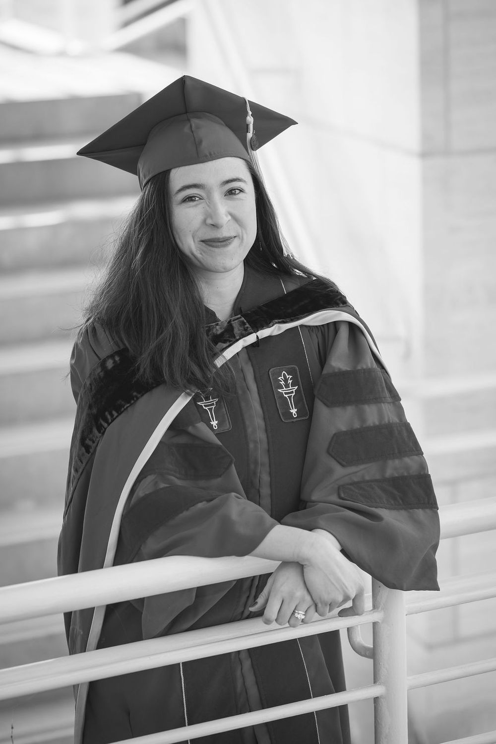Woman in academic graduation cap and gown smiling while leaning on a railing in a black and white photo.