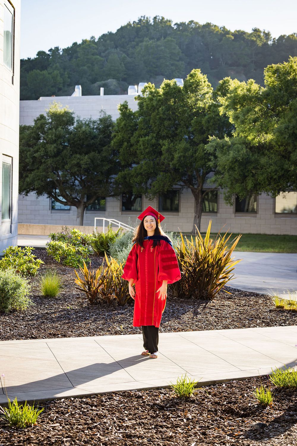 A woman in red graduation cap and gown walks along a campus pathway surrounded by lush greenery and buildings.