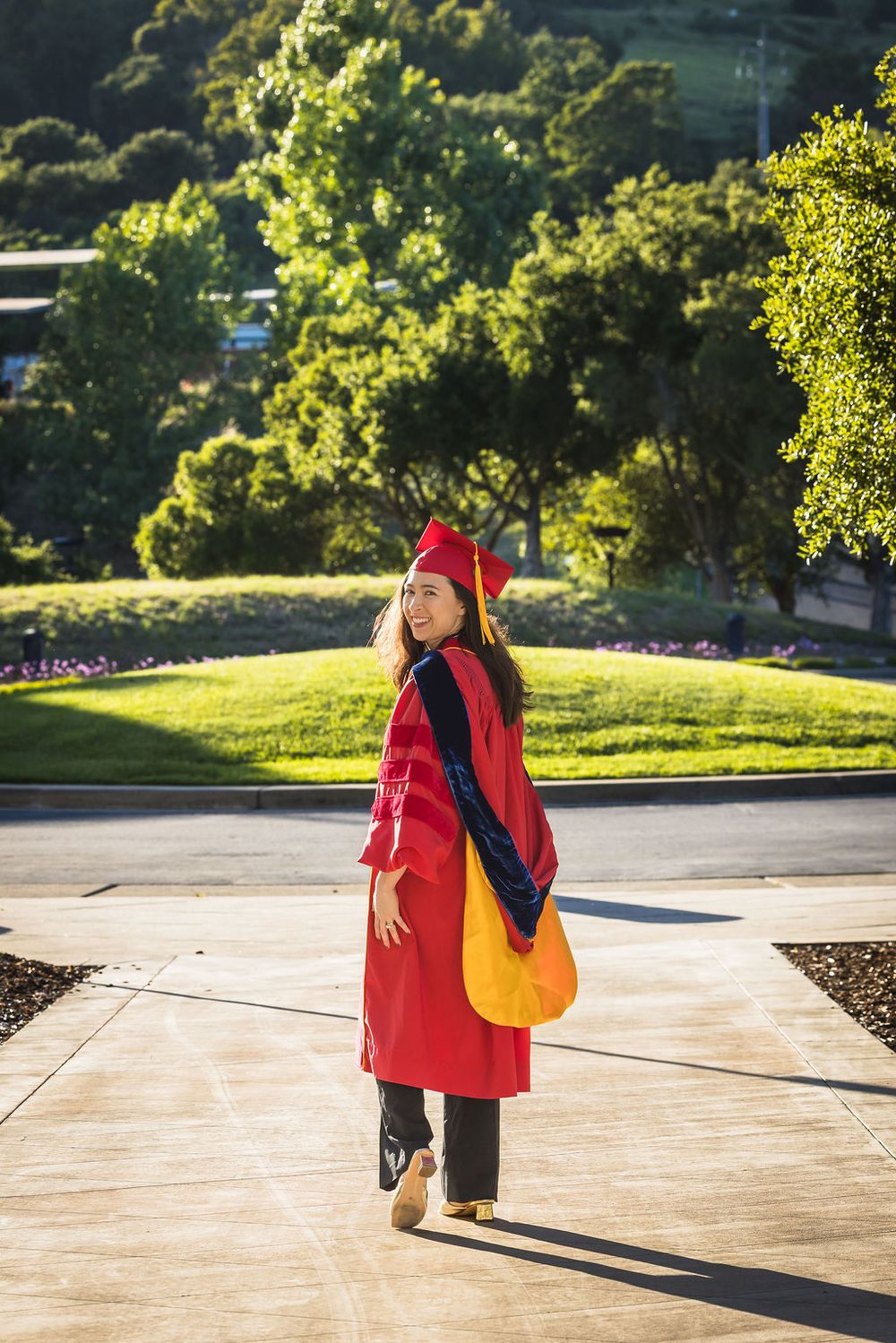 Young woman in red graduation cap and gown smiling outdoors on sunny campus with green trees.