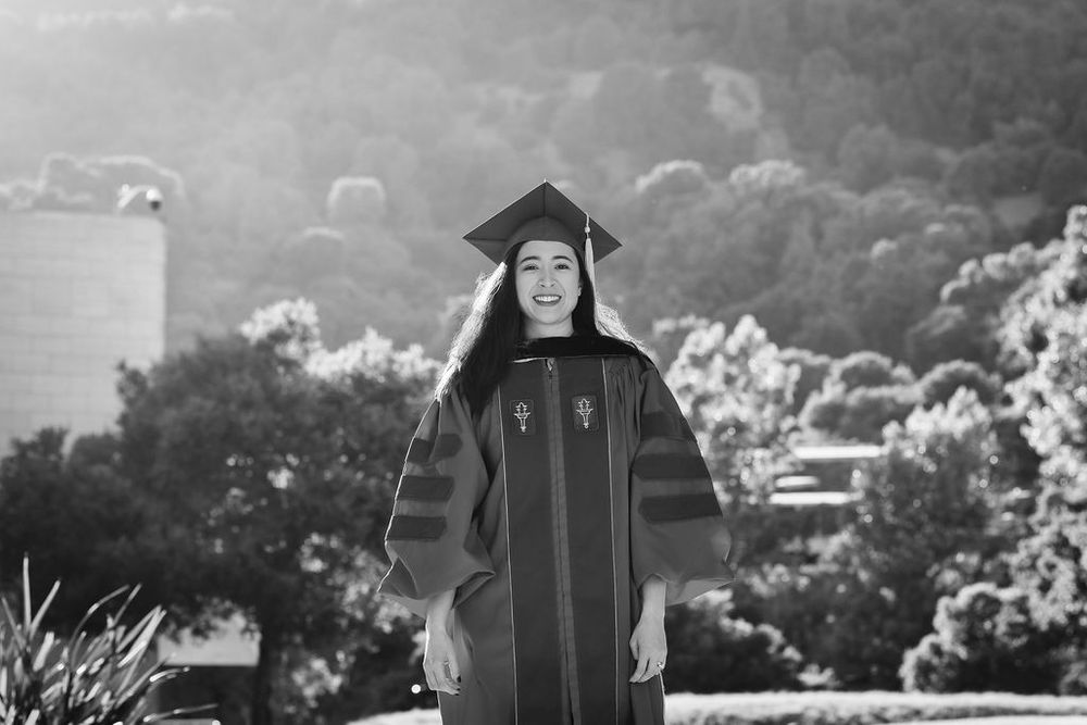 Smiling graduate in cap and gown poses outdoors with tree-covered hills in background, black and white photo.