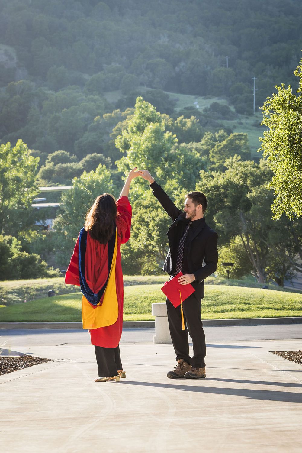 Two graduates in colorful regalia celebrate outdoors with a high-five against a scenic hillside backdrop.