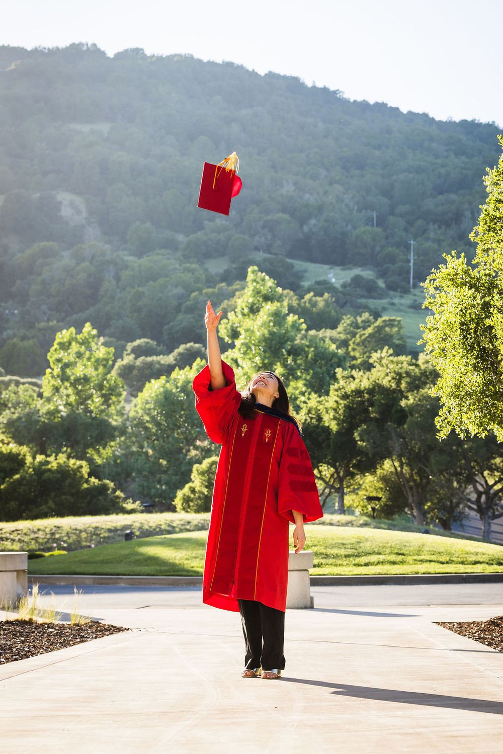 Graduate in red gown tosses cap in air outdoors, celebrating graduation with green hills in background.