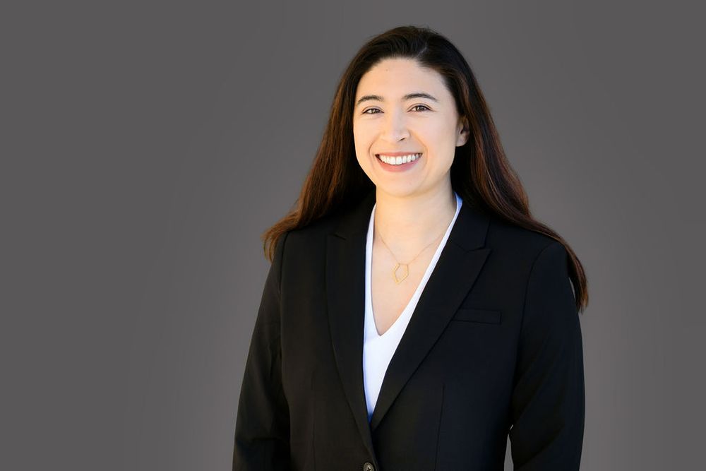 Professional woman in black blazer smiling confidently against gray background in corporate headshot photo.