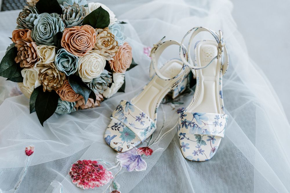 Floral heeled wedding sandals beside a colorful paper flower bouquet on white bridal veil fabric.