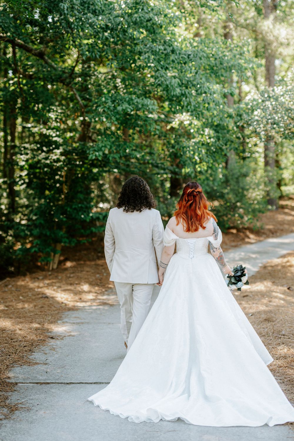 Bride in white gown and groom in beige suit walk hand-in-hand down a tree-lined path on their wedding day.
