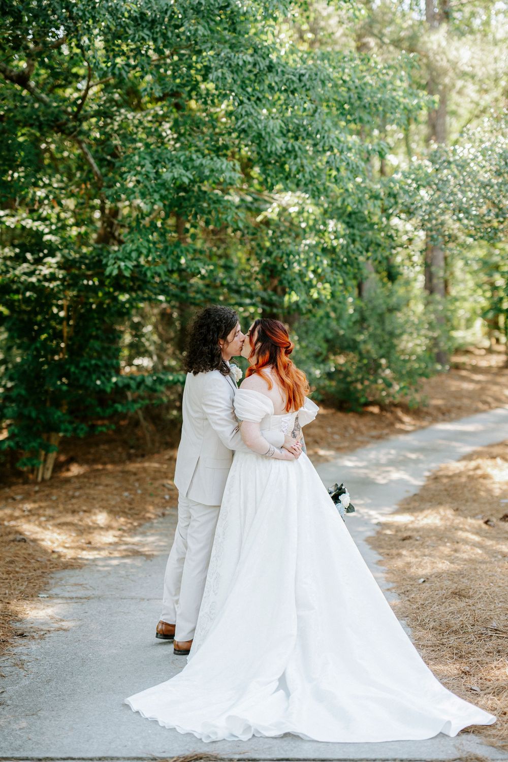 Bride and groom share a romantic kiss on a tree-lined path, bride wearing flowing white off-shoulder gown with long train.