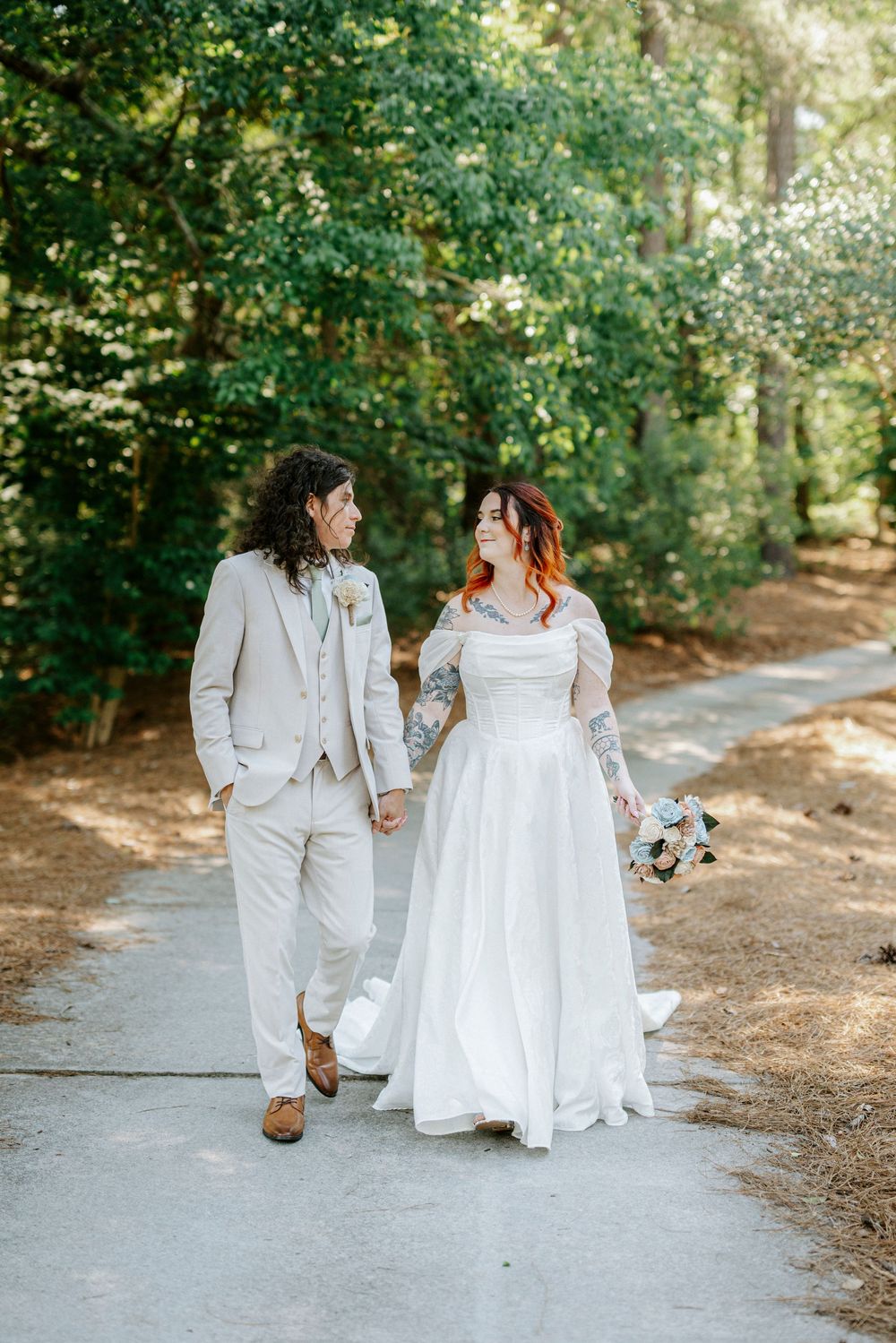 Bride and groom walk hand-in-hand along a sunlit forest path, bride in white gown holding bouquet, groom in beige suit.