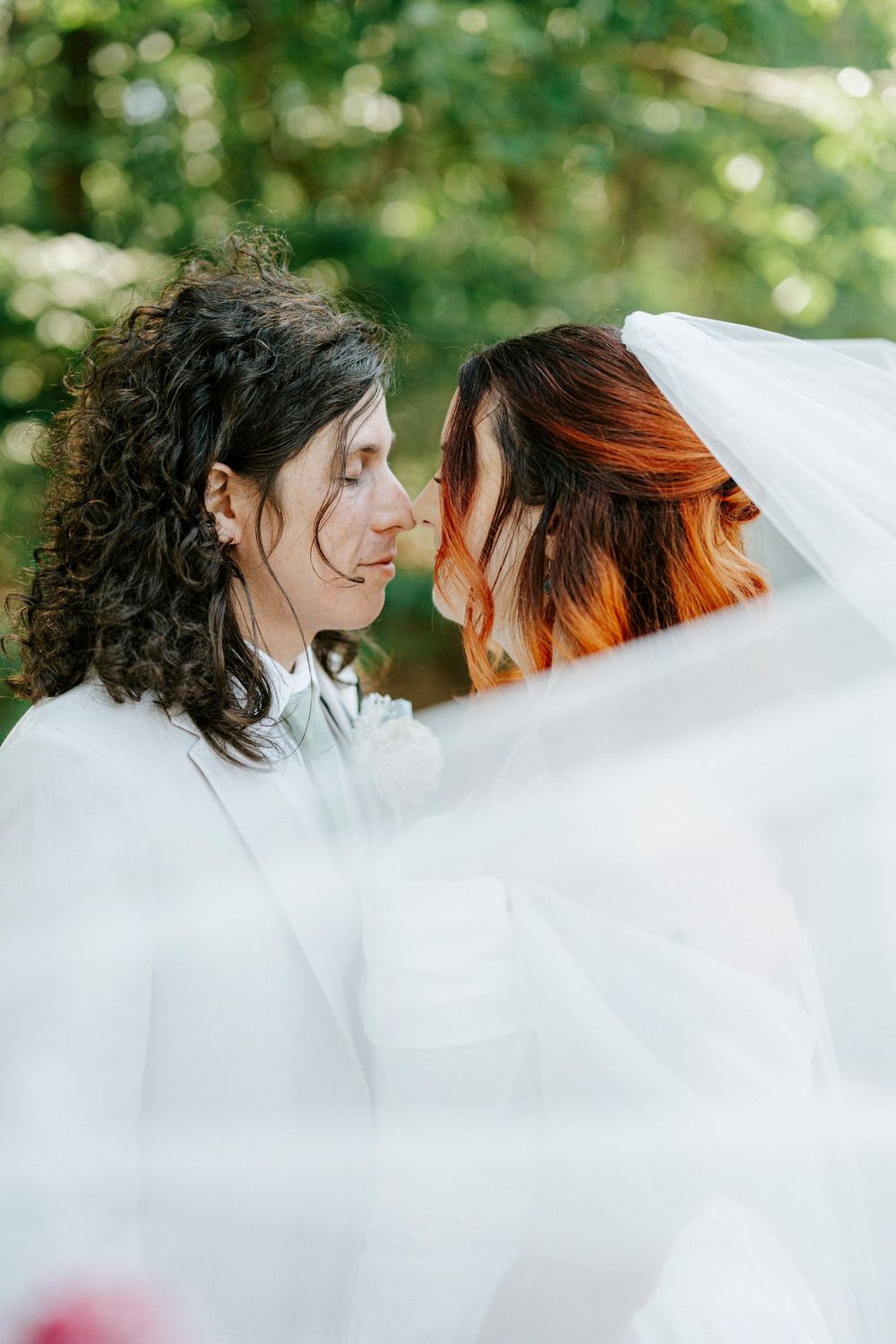 Two brides share an intimate moment under a flowing veil outdoors, surrounded by lush green trees.
