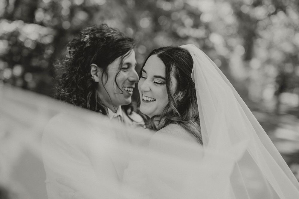 Black and white wedding photo of smiling bride and groom sharing a joyful moment under a flowing veil outdoors.