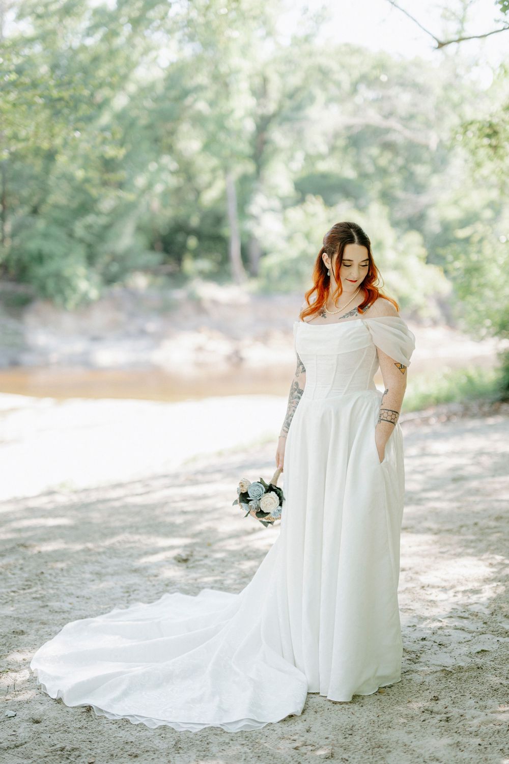 Bride in off-shoulder white gown with train holds bouquet outdoors near a sunlit creek.