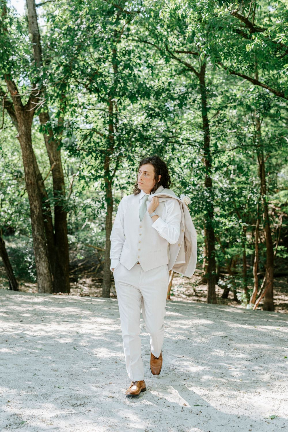 Groom in beige suit carries jacket over shoulder while walking through a sun-dappled forest clearing.