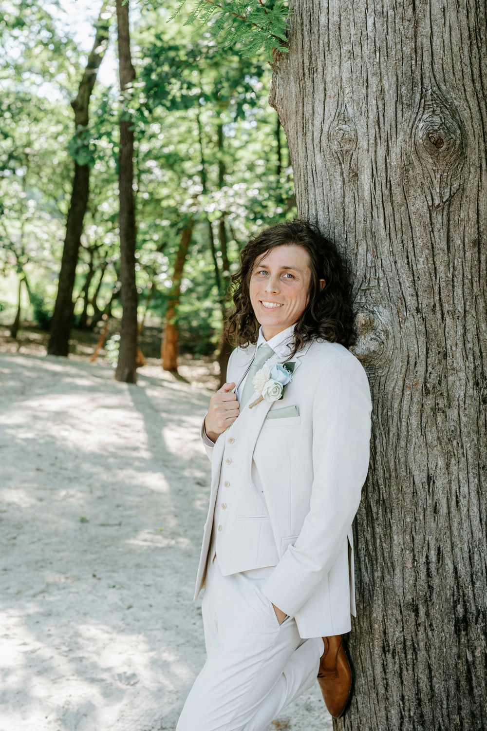 Person in white suit leaning against tree trunk in forest, smiling with boutonniere, wedding attire.