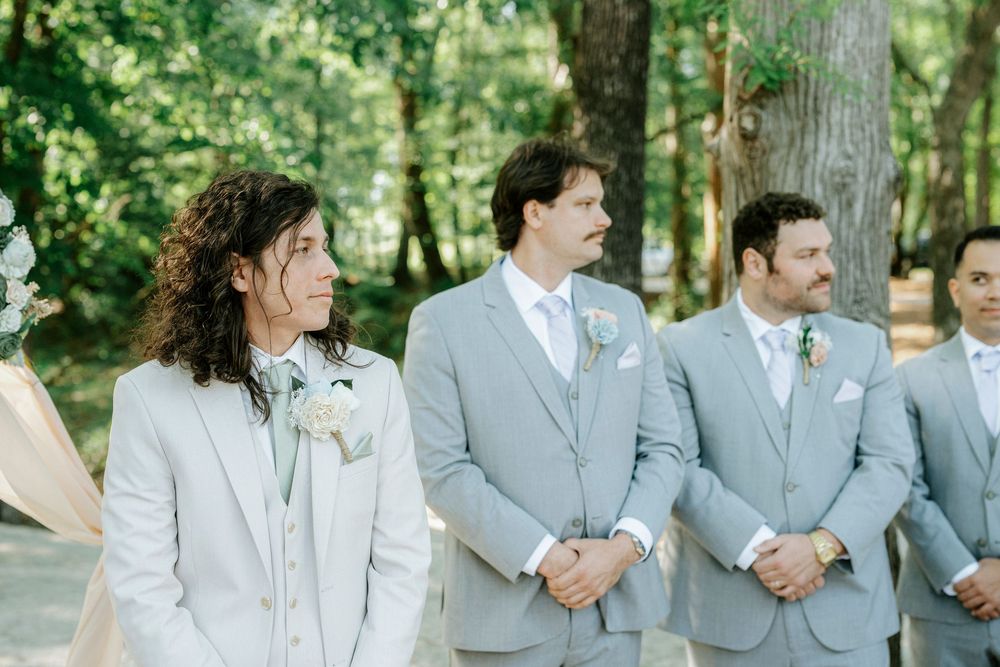 Groomsmen in light gray suits stand at outdoor woodland wedding ceremony.