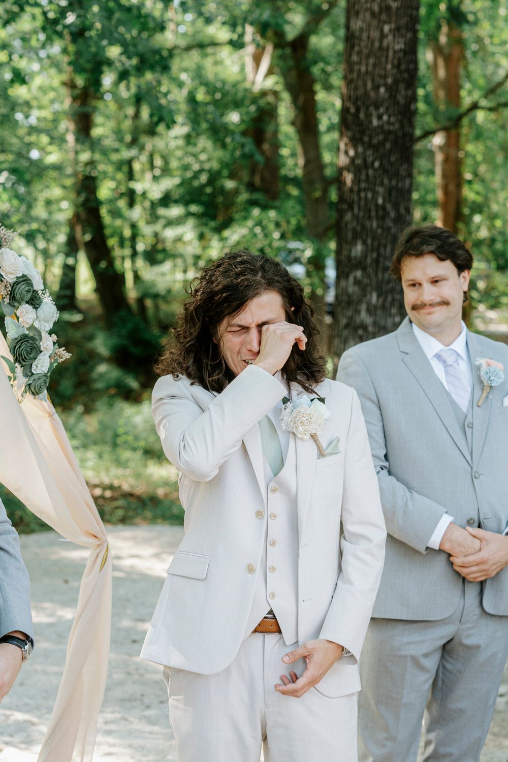 Emotional groom wiping tears at outdoor wedding ceremony in forest with groomsman in gray suit behind him.