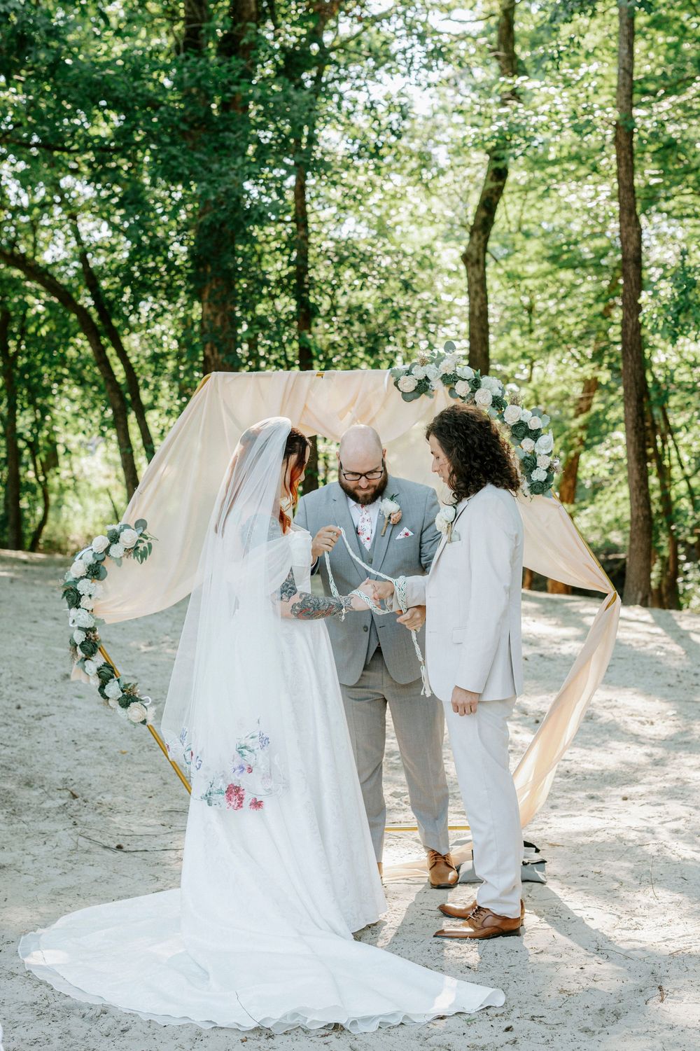 Bride and groom perform handfasting ceremony under floral arch on sandy woodland clearing with officiant.