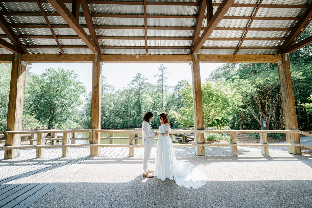 Two brides stand together under a wooden pavilion surrounded by lush greenery on their wedding day.