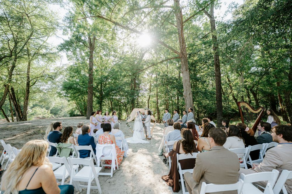 Outdoor woodland wedding ceremony with guests seated in white chairs.