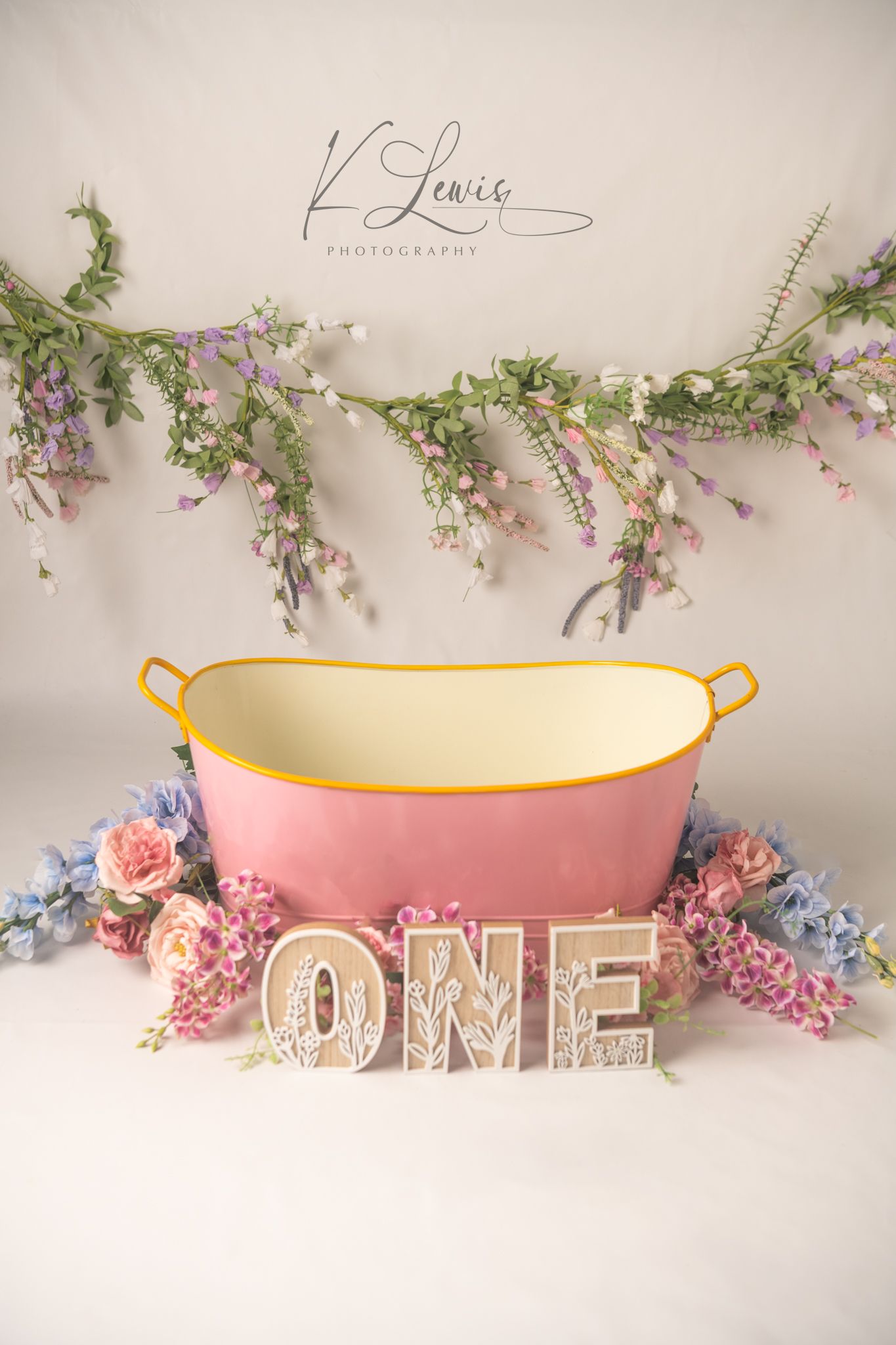 Pink enamel tub with yellow trim surrounded by pastel flowers and ONE letters for a first birthday cake smash photo session.