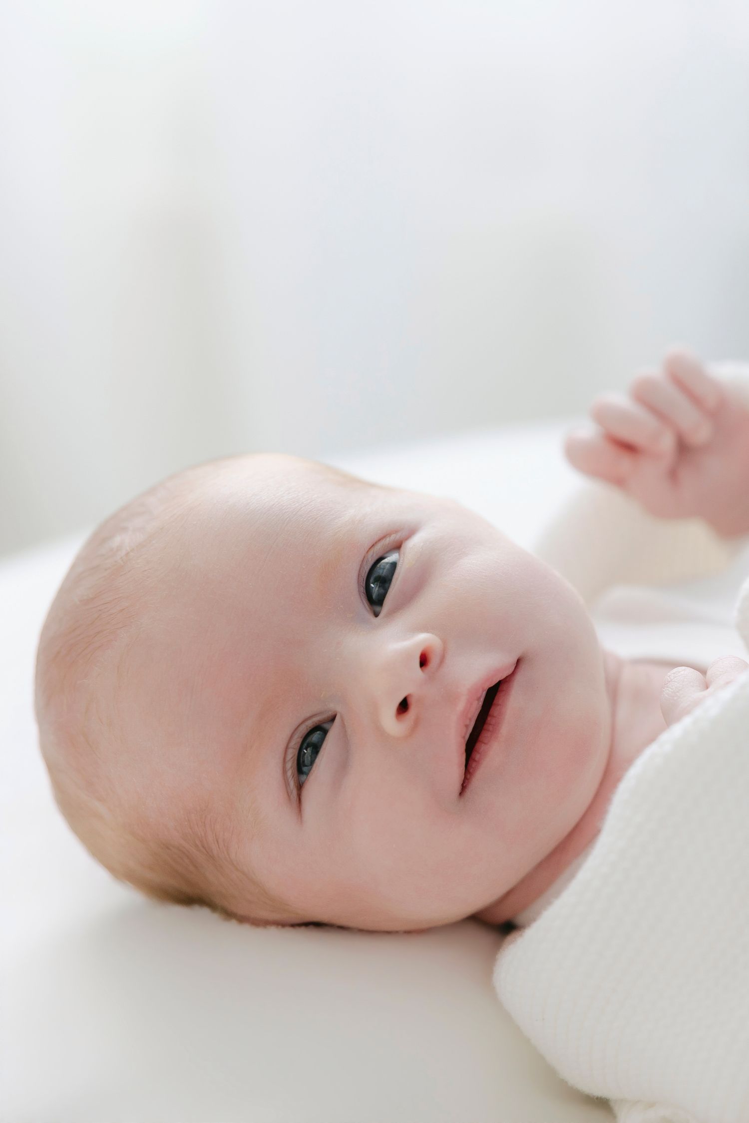 A peaceful newborn baby with blue eyes lying on a white surface, gently raising a tiny fist.