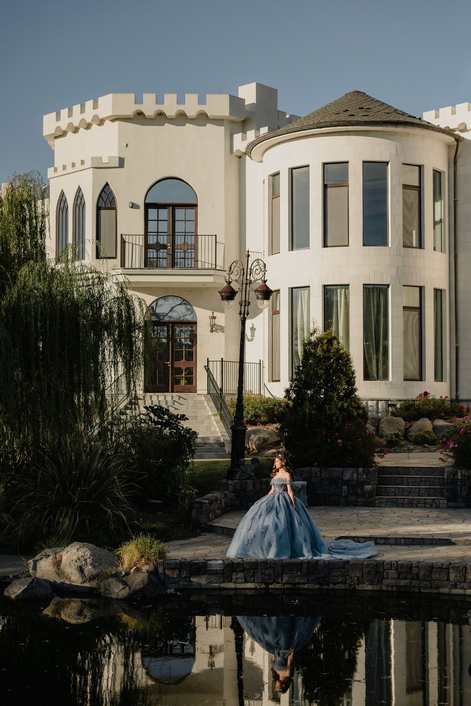 Woman in blue ball gown poses by a pond in front of a white castle-like mansion on a sunny day.