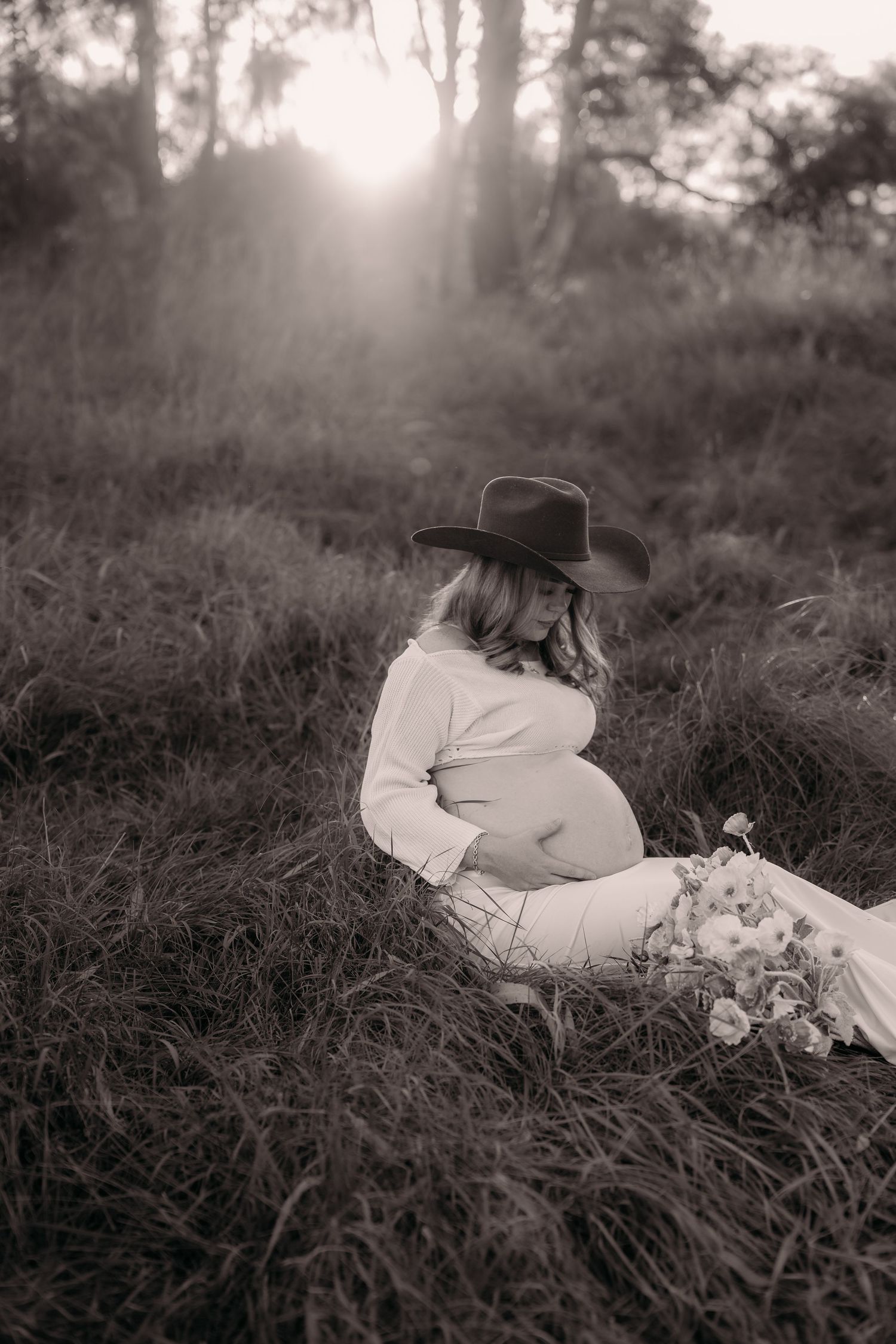 Pregnant woman in cowboy hat sitting in grass field at sunset, black and white maternity photo with wildflowers.
