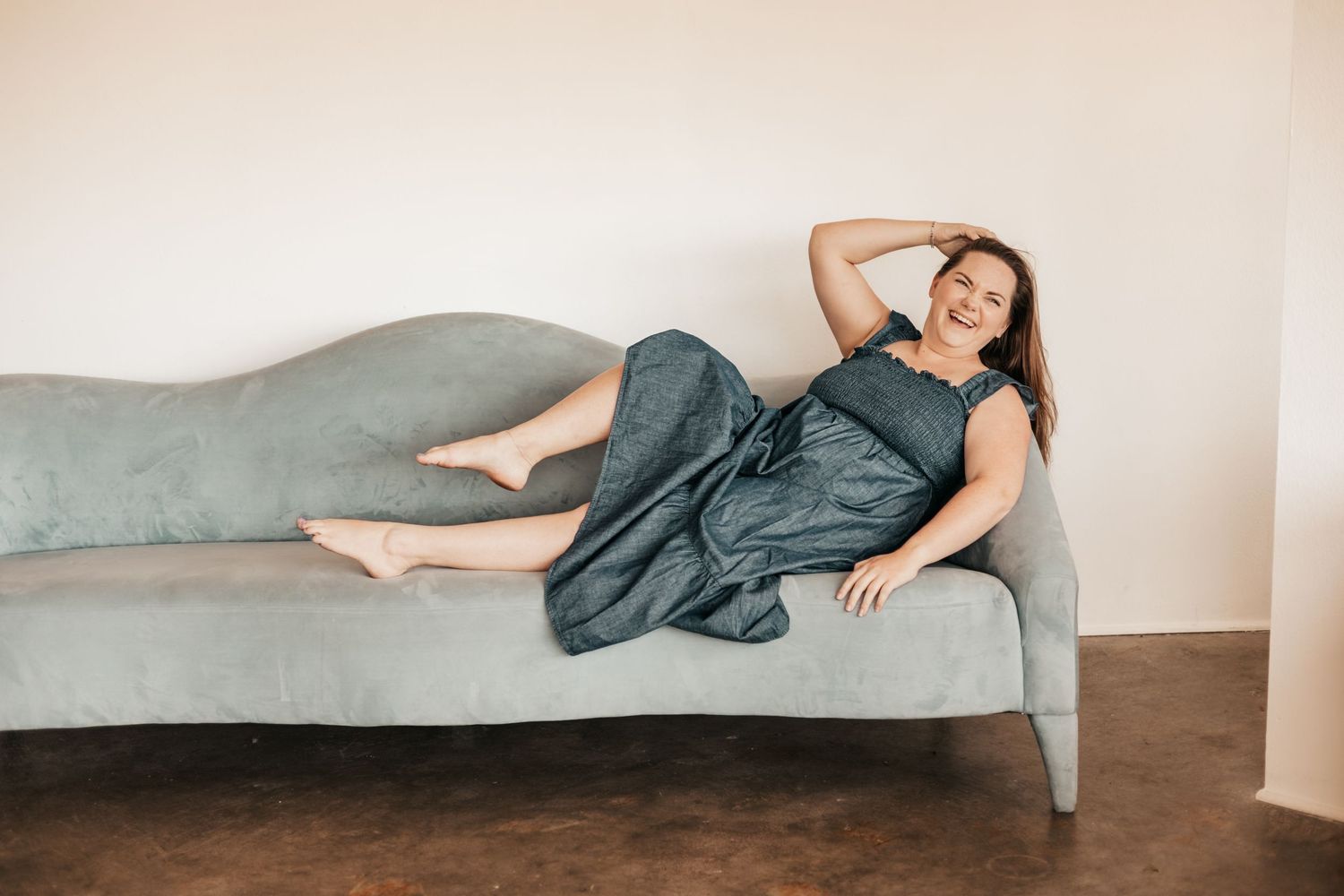 Woman in teal dress lounging on a gray chaise sofa, smiling and relaxed in a bright minimalist room | Photography Business Coach