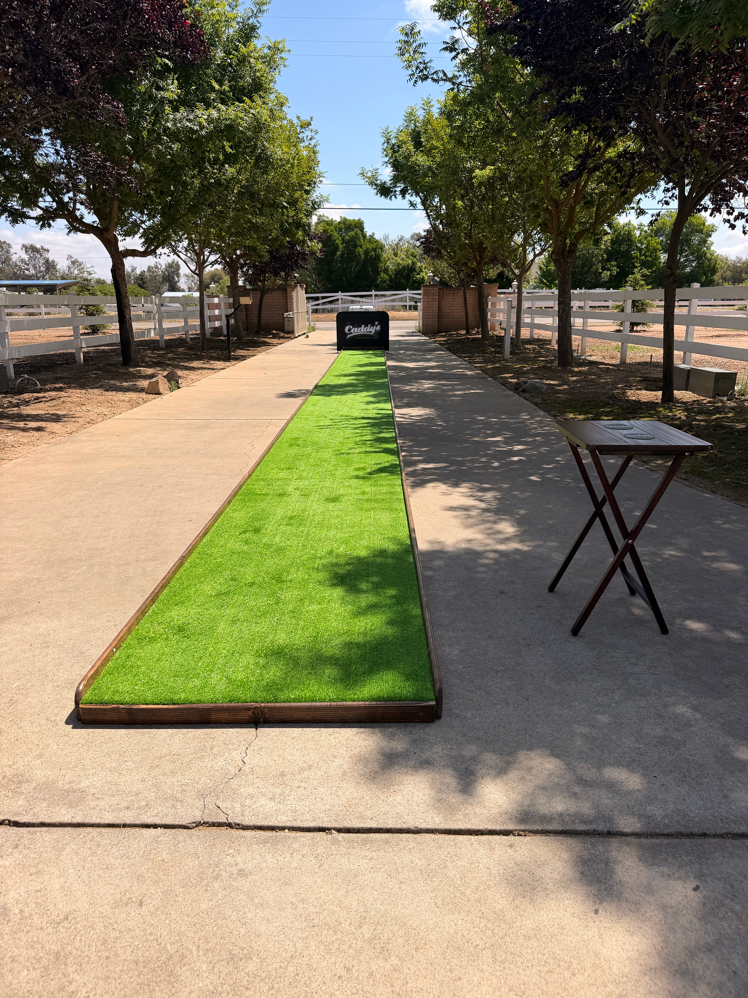Outdoor putting green on a concrete path lined with trees and white fences, with a folding table nearby.
