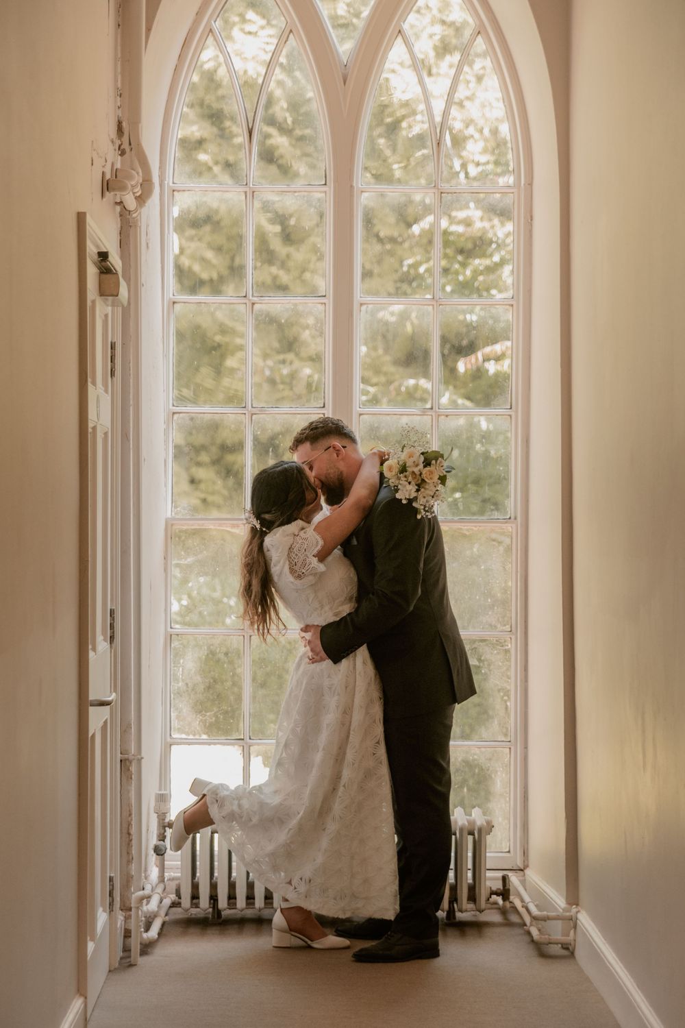 Bride and groom sharing a kiss in front of an arched Gothic window indoors.