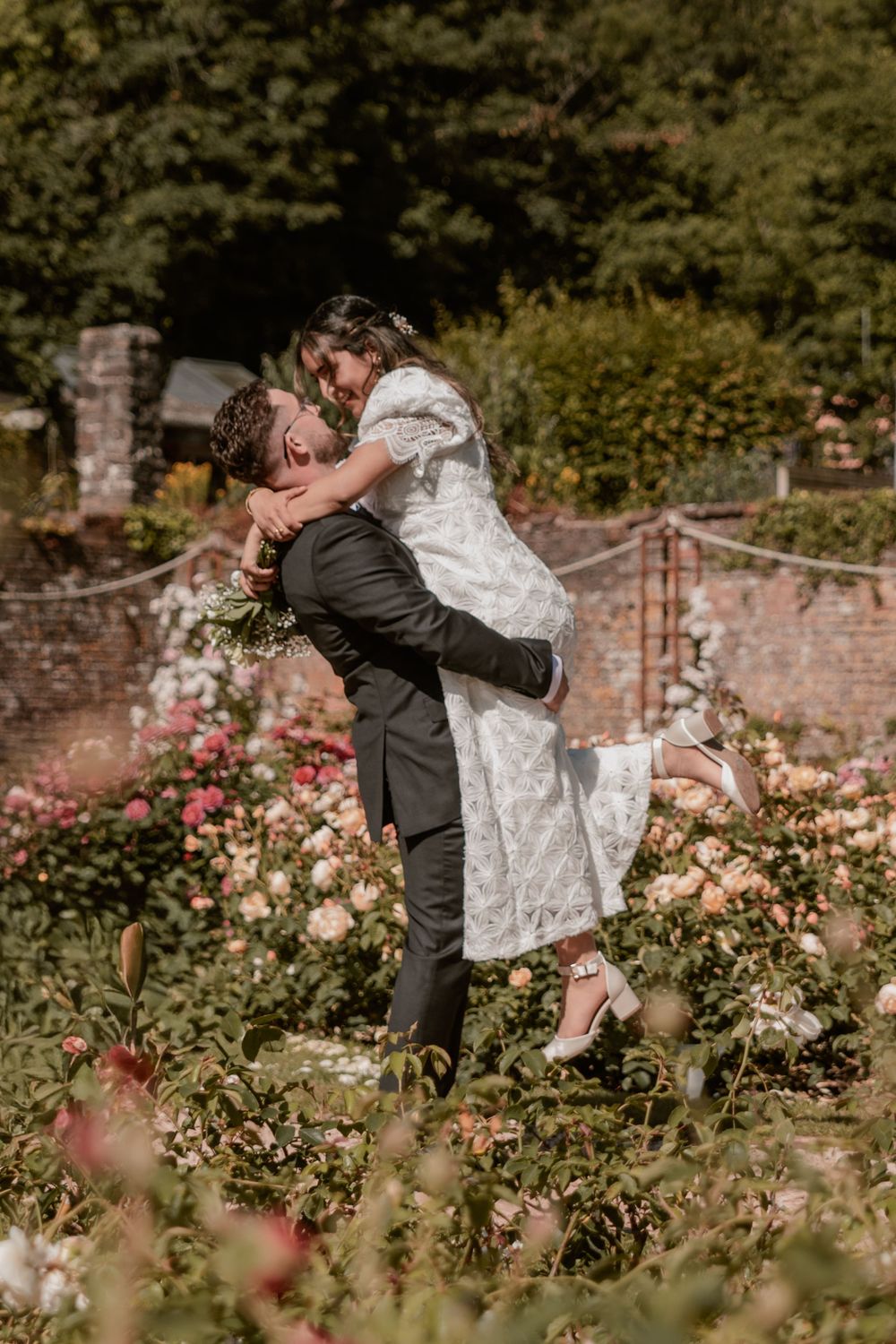 Groom lifting bride in lace dress among blooming roses in a garden wedding photo.
