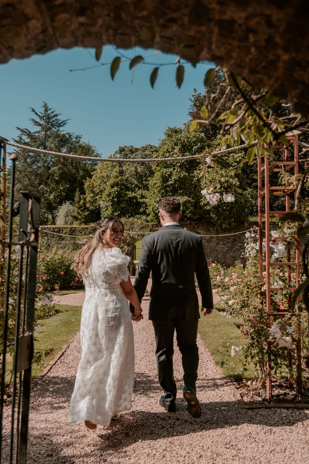 Bride and groom walking hand in hand through a garden gate on their wedding day.