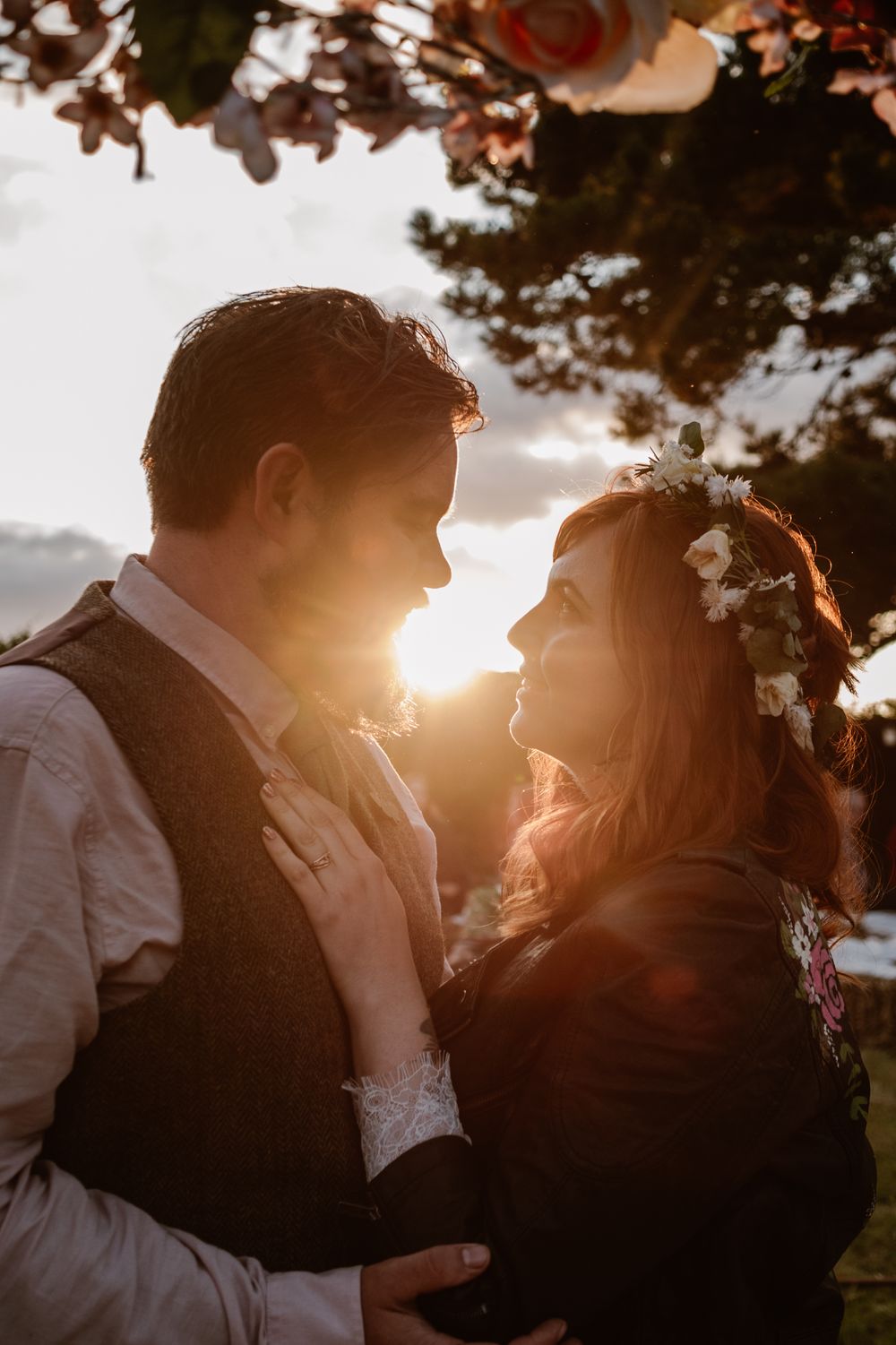 Couple embracing at golden sunset, bride wearing floral crown, warm backlit romantic wedding portrait outdoors.