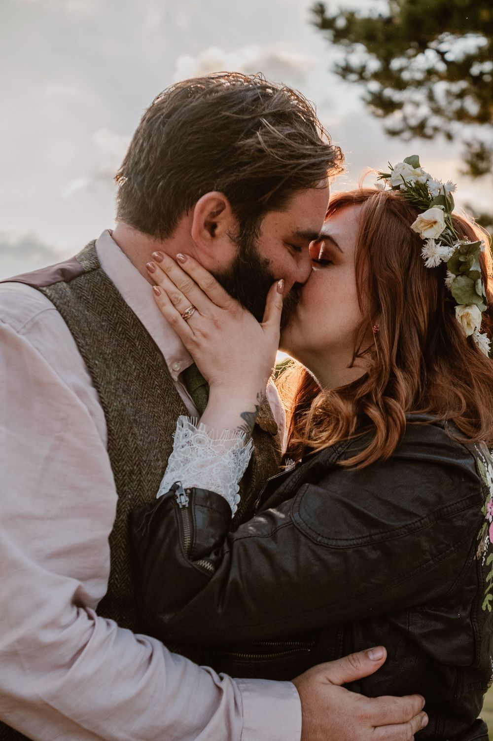 Couple sharing a romantic kiss outdoors, bride wearing floral crown and leather jacket, groom in vest at golden hour.