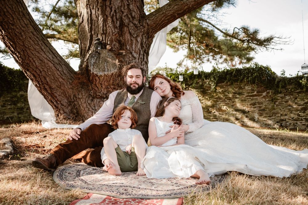 A happy family of four poses under a large tree in a rustic outdoor wedding setting with the couple in formal attire.