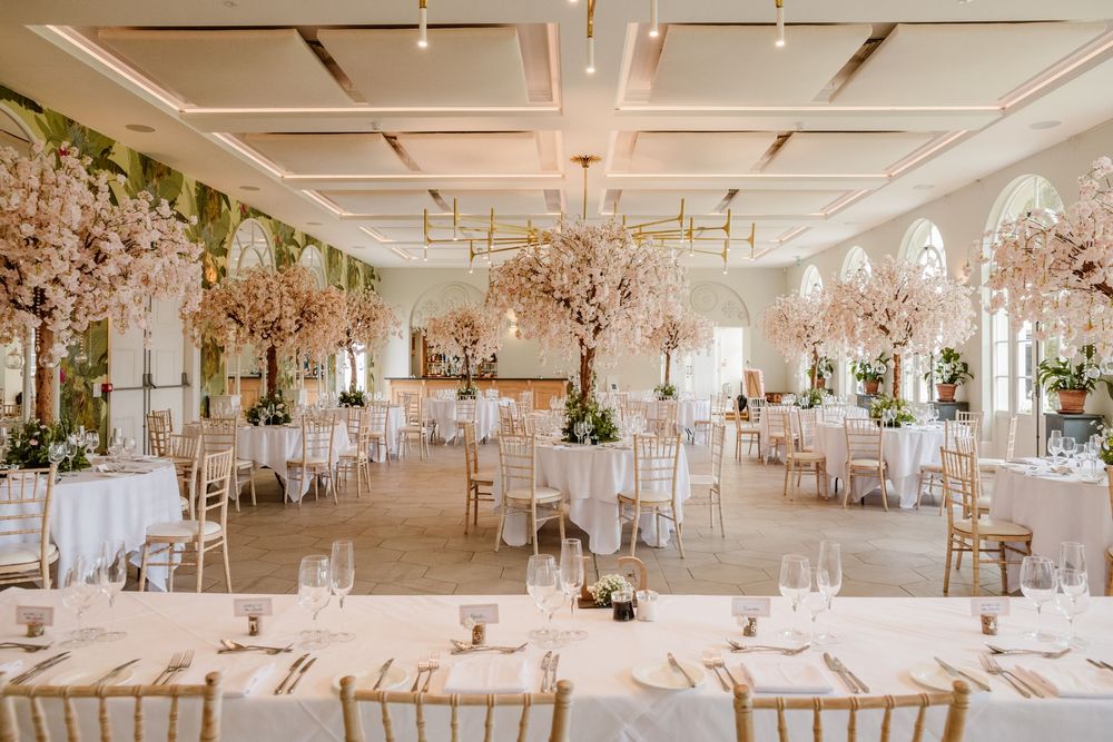 Elegant wedding reception hall with blush floral arrangements, gold chiavari chairs, and white draped tables in a bright ballroom.