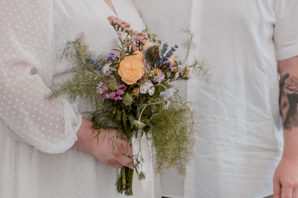 Bride holding a wildflower wedding bouquet with peach rose, blue and pink blooms, and greenery in white dress.